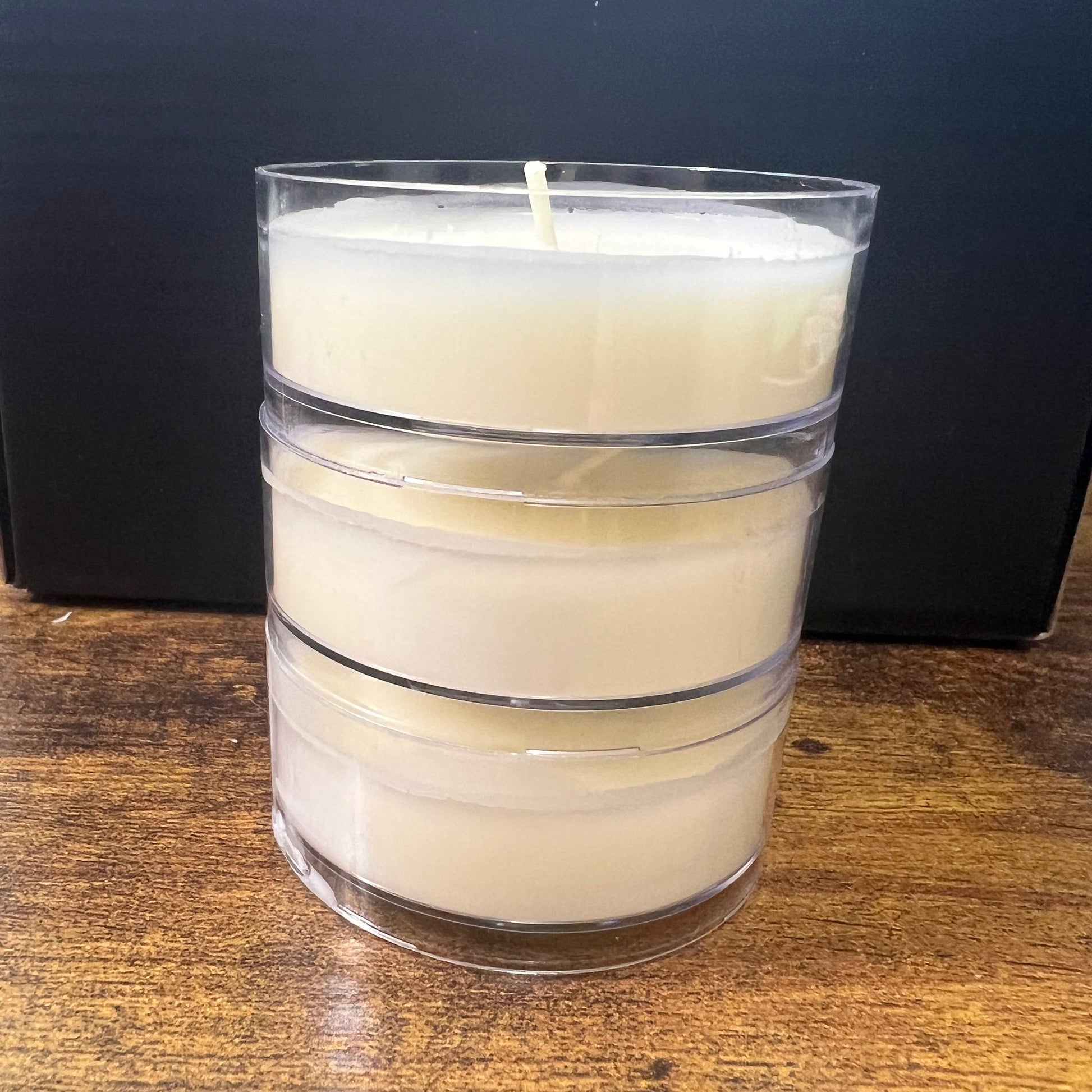 Wide tea light candles shown standing upright on a dark surface next to a wooden tabletop.