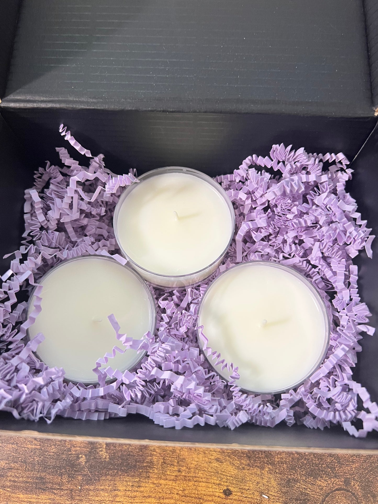 Three large tealight candles in clear holders, nestled in purple shredded paper filler inside a black gift box.