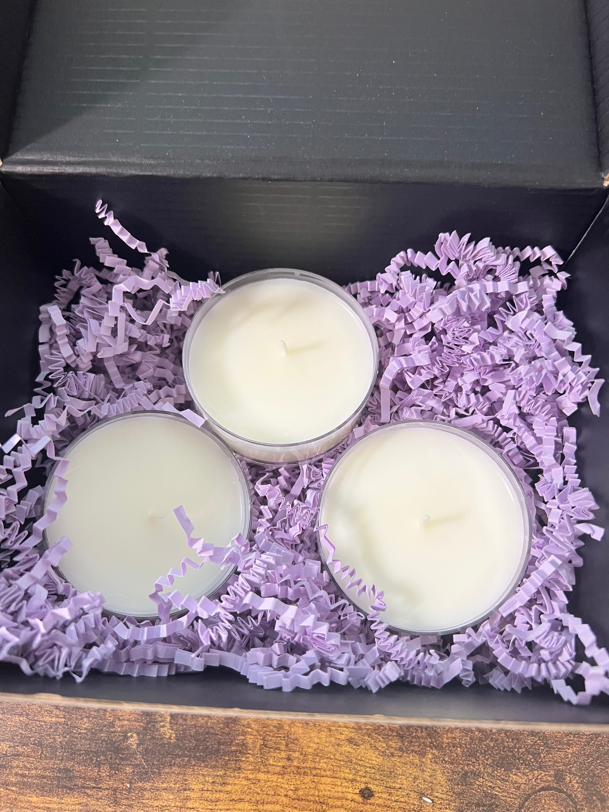 Three large tealight candles in clear holders, nestled in purple shredded paper filler inside a black gift box.