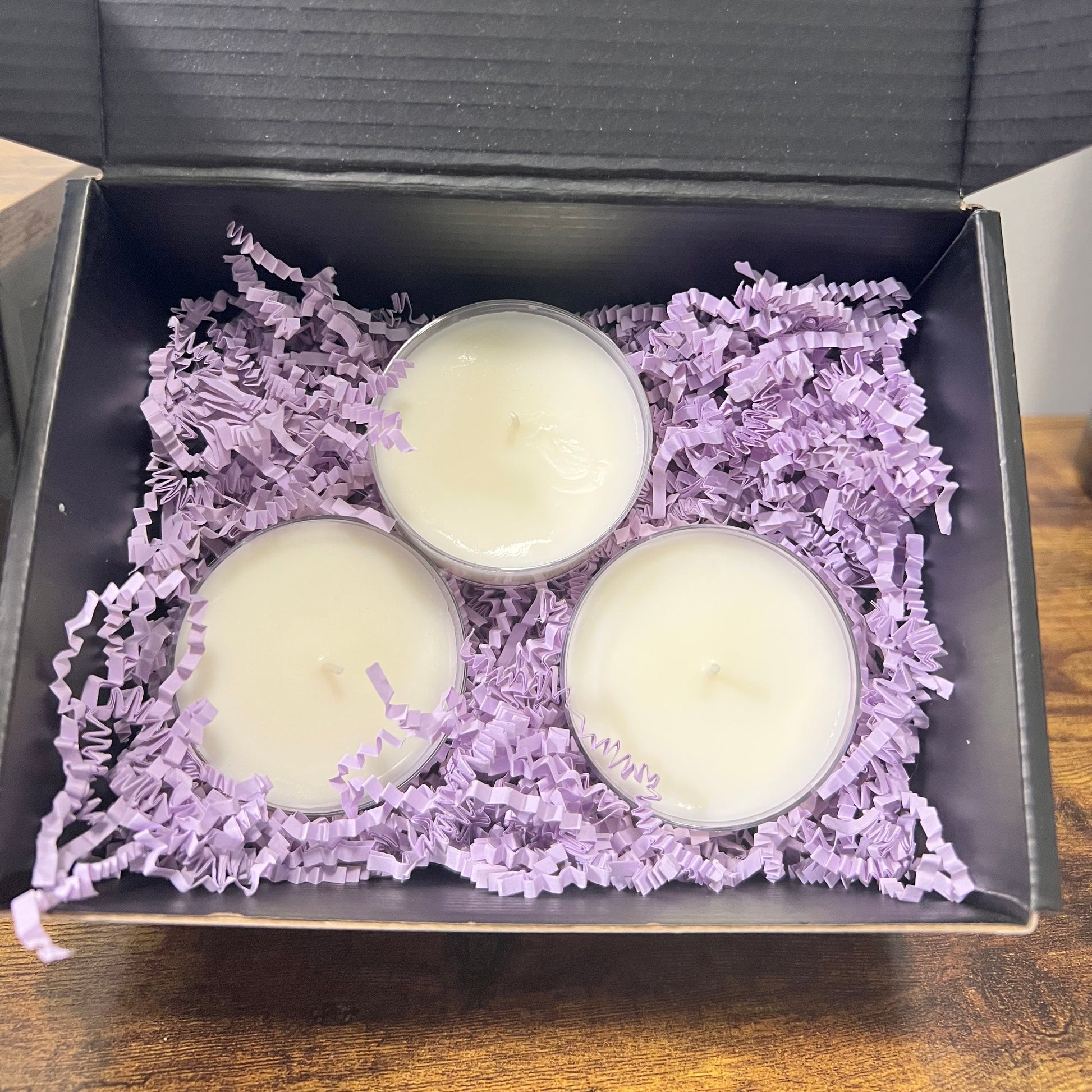 Three small white candles in clear cups nestled in lavender purple shredded paper inside an open  gift box, placed on a wooden surface.