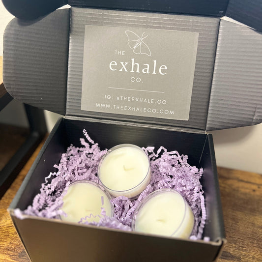 Three large tea light candles in clear holders, nestled in lavender crinkle paper inside an open black gift box. The inside lid features a gray label for 'The exhale co'.