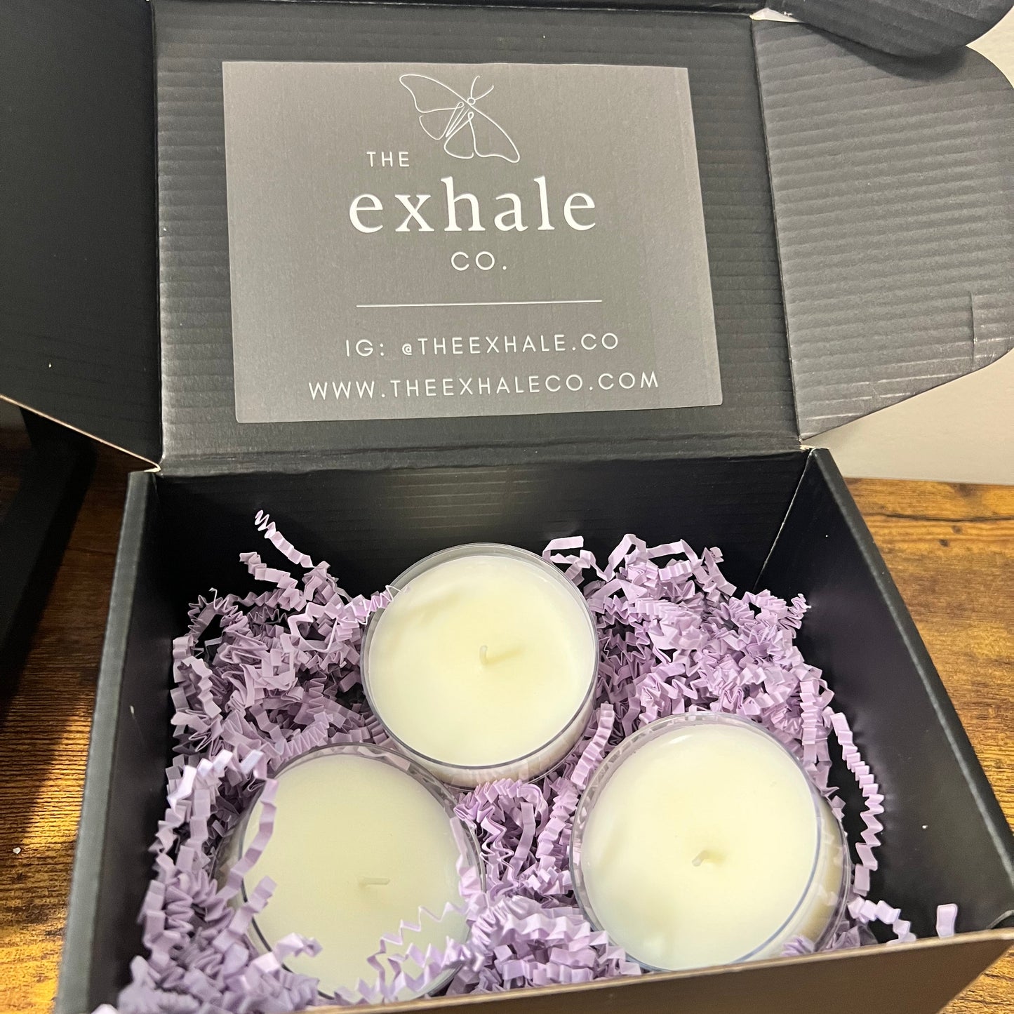 Three large tea light candles nestled in light purple shredded paper inside an open black gift box. 